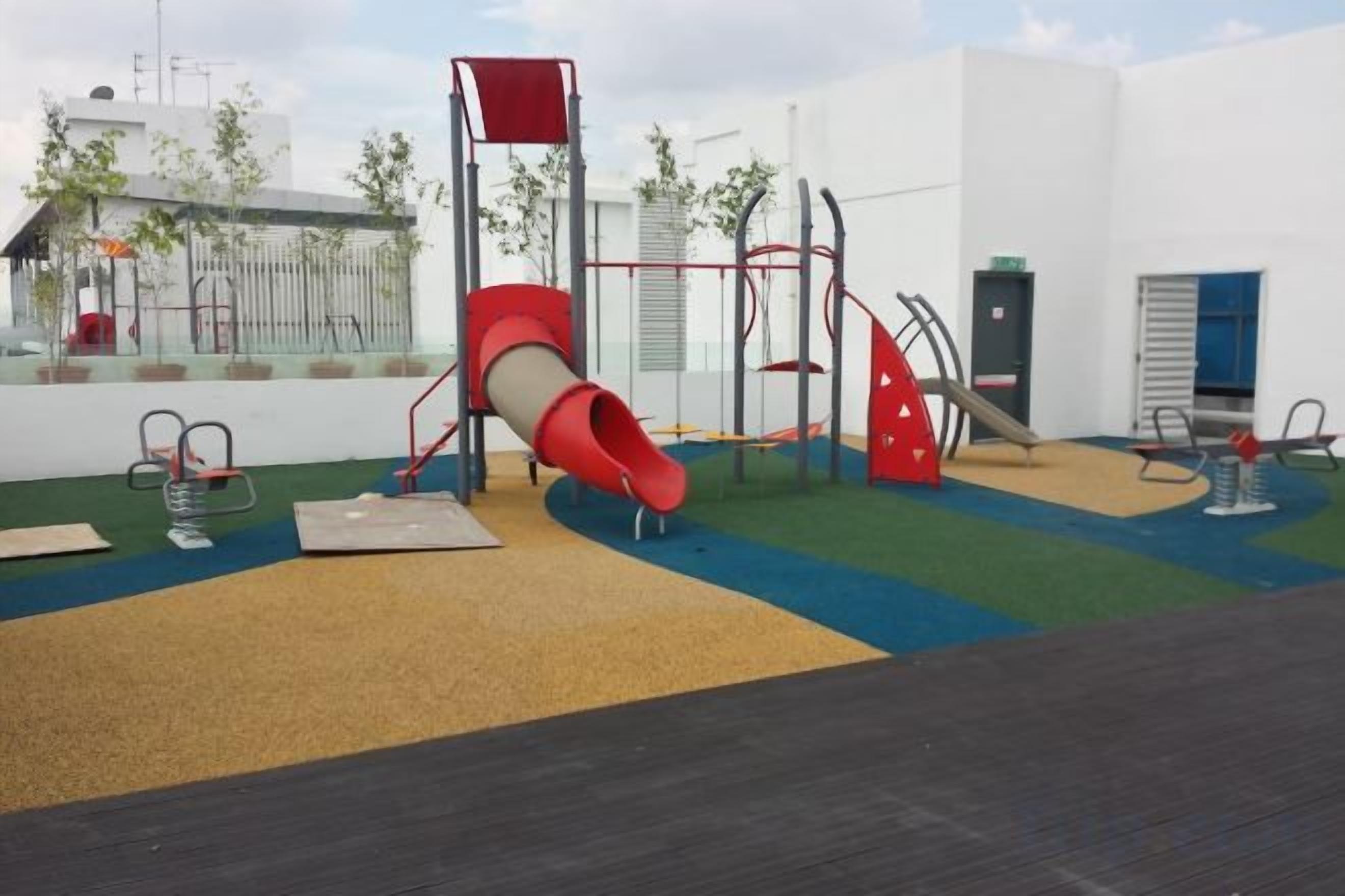 A playground with a slide and climbing structures, surrounded by a fenced area and a building in the background.