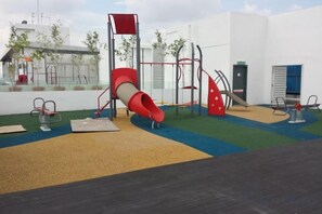 Een speeltuin met een glijbaan en klimconstructies, omringd door een hek en een gebouw op de achtergrond.