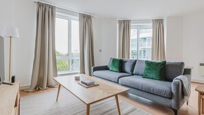 A modern living room with a grey sofa, green cushions, a wooden coffee table, and a view of buildings through large windows. - Sleek Southwark 2BR, Furniture & Decor Included, by Blueground (London)