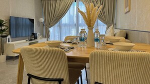 Une salle à manger moderne avec une table en bois, des chaises recouvertes d’un tissu beige, un vase contenant des plumes d’herbe des pampas, des bouteilles d’eau et un téléviseur fixé au mur.