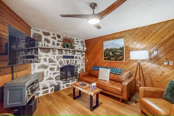 A cozy living room with a stone fireplace, wooden walls, a leather sofa, and a television.