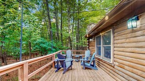 A wooden deck with Adirondack chairs and a table, surrounded by a forest.