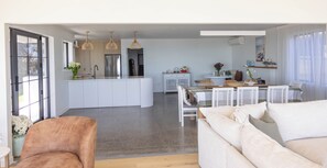 A modern kitchen with white cabinetry, a central island, pendant lighting, and a dining area with a table and chairs. - Beachfront Luxury Retreat & Sauna with Sea Views (New Plymouth)