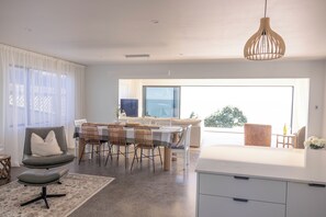 A modern living room with a dining area, a sofa, a table with chairs, and a view to the outside. - Beachfront Luxury Retreat & Sauna with Sea Views (New Plymouth)