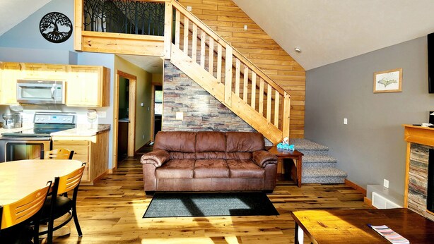 A spacious living area with a brown leather couch, wooden flooring, a kitchen with a microwave, and a wooden staircase. - Catchin Crickets near Pictured Rocks and ATV Trails!  (Shingleton)