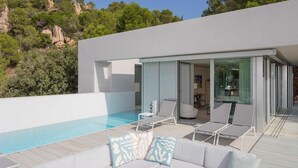 Ein moderner Poolbereich im Freien mit einem weißen Infinity-Pool, einer weißen Couch mit Kissen sowie einer Glastür, die auf eine überdachte Terrasse führt.