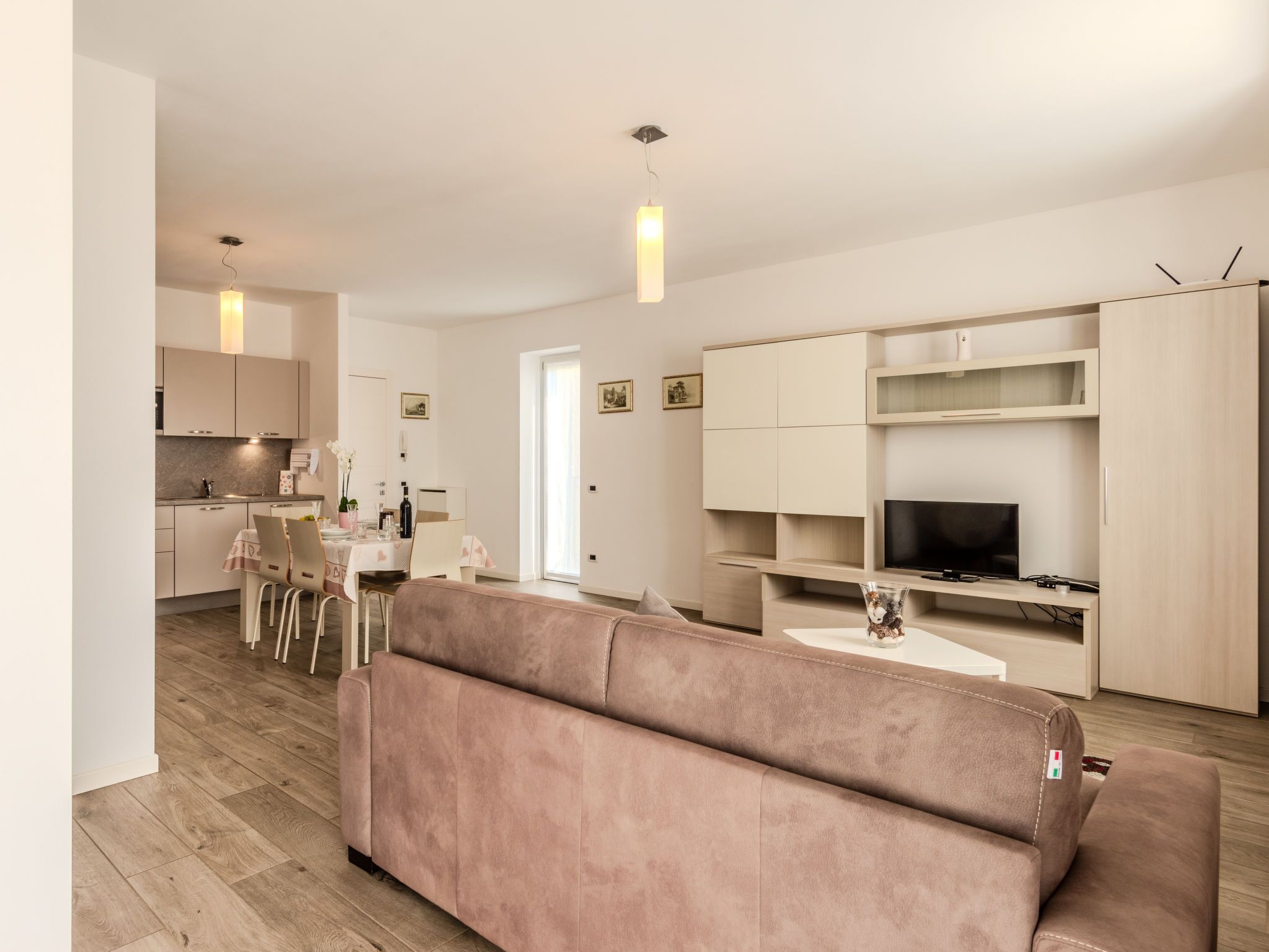 A modern living room with a beige sofa, a dining area with a table and chairs, and a kitchen with cabinets and a TV.