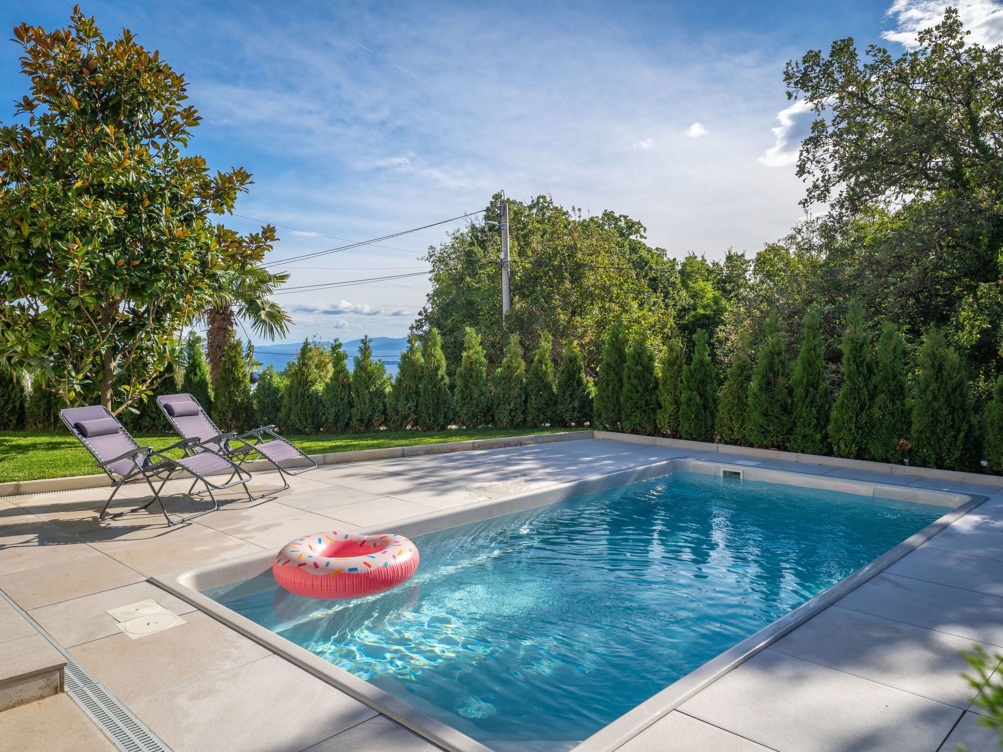 A rectangular swimming pool with a pink inflatable ring, surrounded by a tiled deck and two lounge chairs.