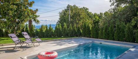 A rectangular swimming pool with a pink inflatable ring, surrounded by a tiled deck and two lounge chairs.