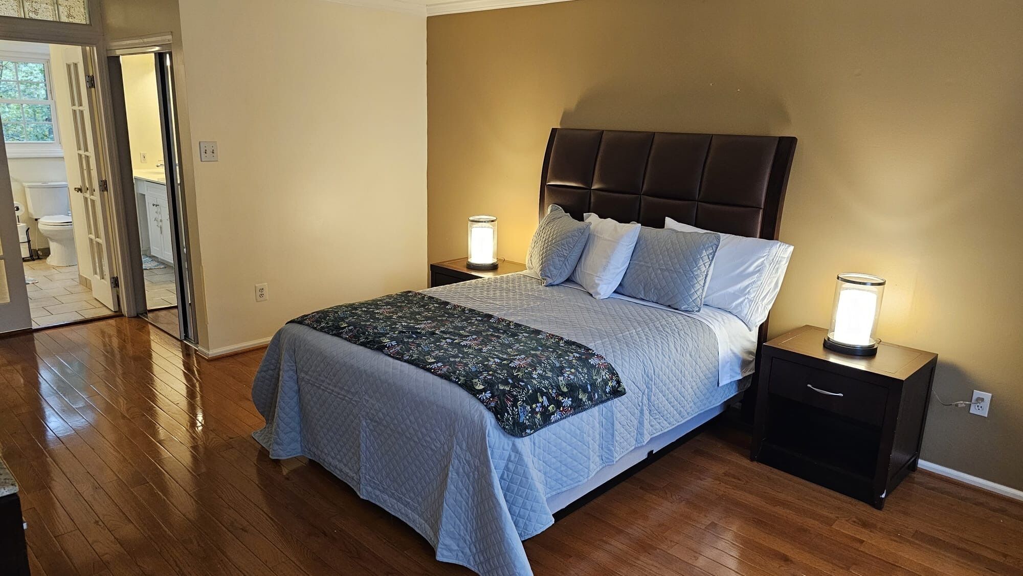 Una camera da letto con un letto grande, due comodini con lampade, pavimento in legno e una porta che dà su un'altra stanza.