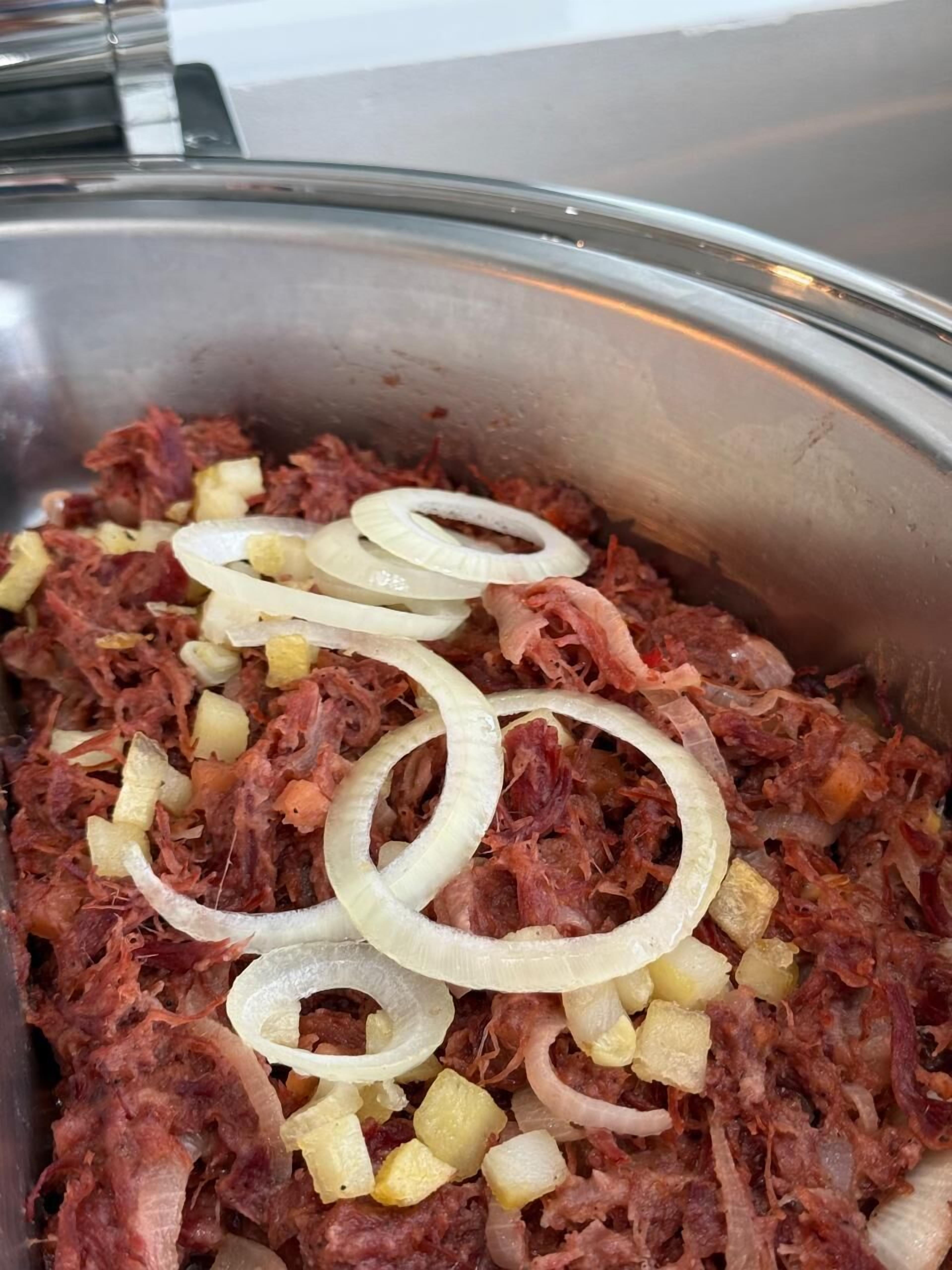 A large metal container filled with shredded meat, diced potatoes, and sliced onions.