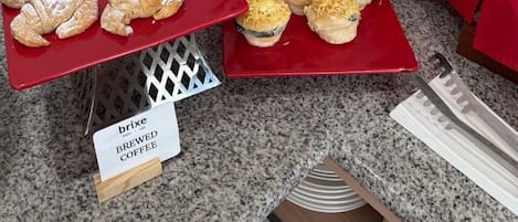 Una mesa de buffet con diversos productos de pastelería, como cruasanes y panecillos, y un letrero que indica café recién hecho.