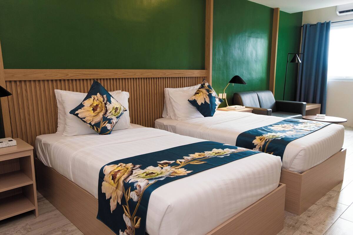 Habitación de hotel con dos camas, cada una con cubrecama y almohadas estampadas con motivos florales, cabecero de madera, escritorio con lámpara y vista al exterior a través de una ventana con cortinas.