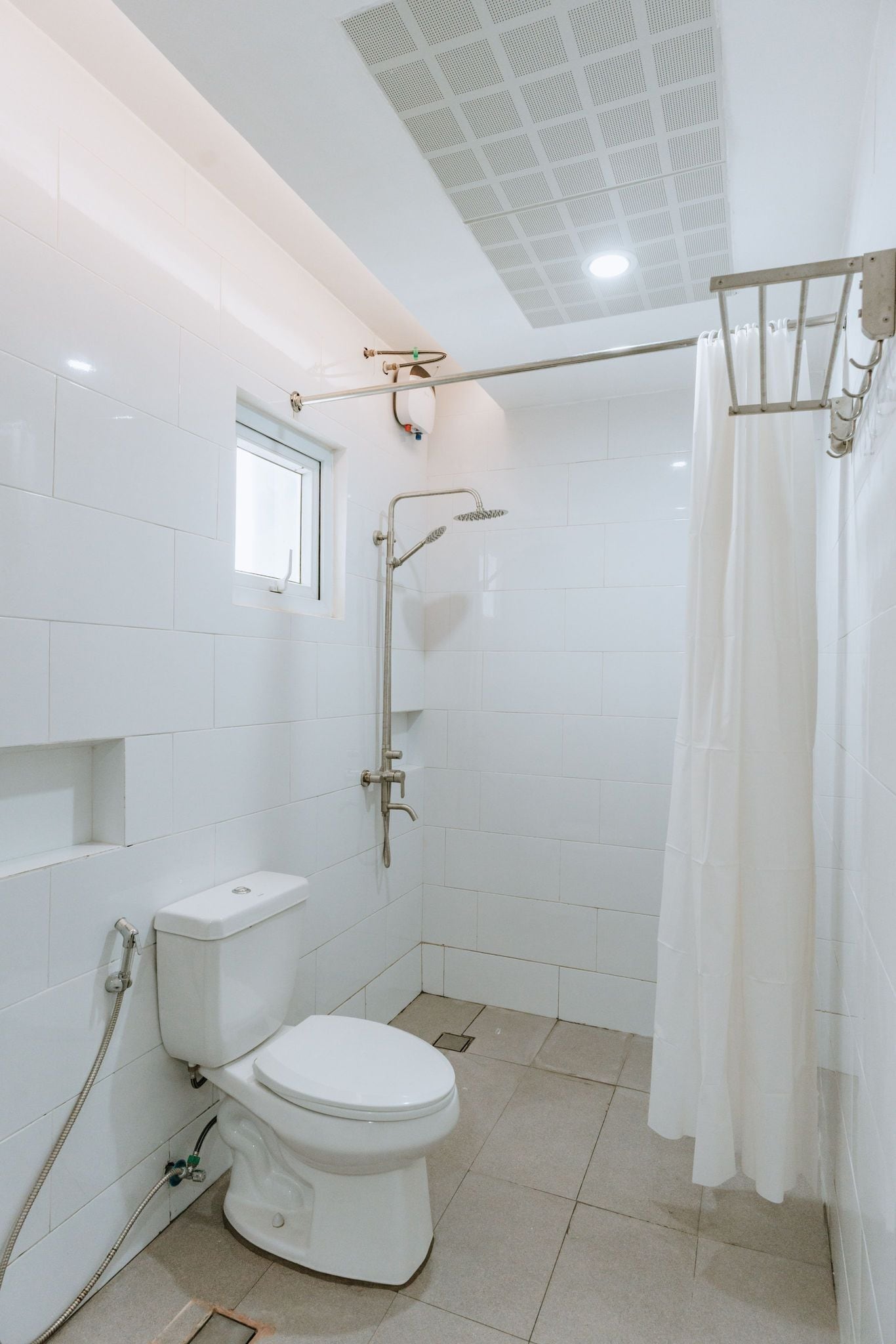 A modern bathroom with white tiles, a toilet, a shower with a glass door, and a window above the toilet.