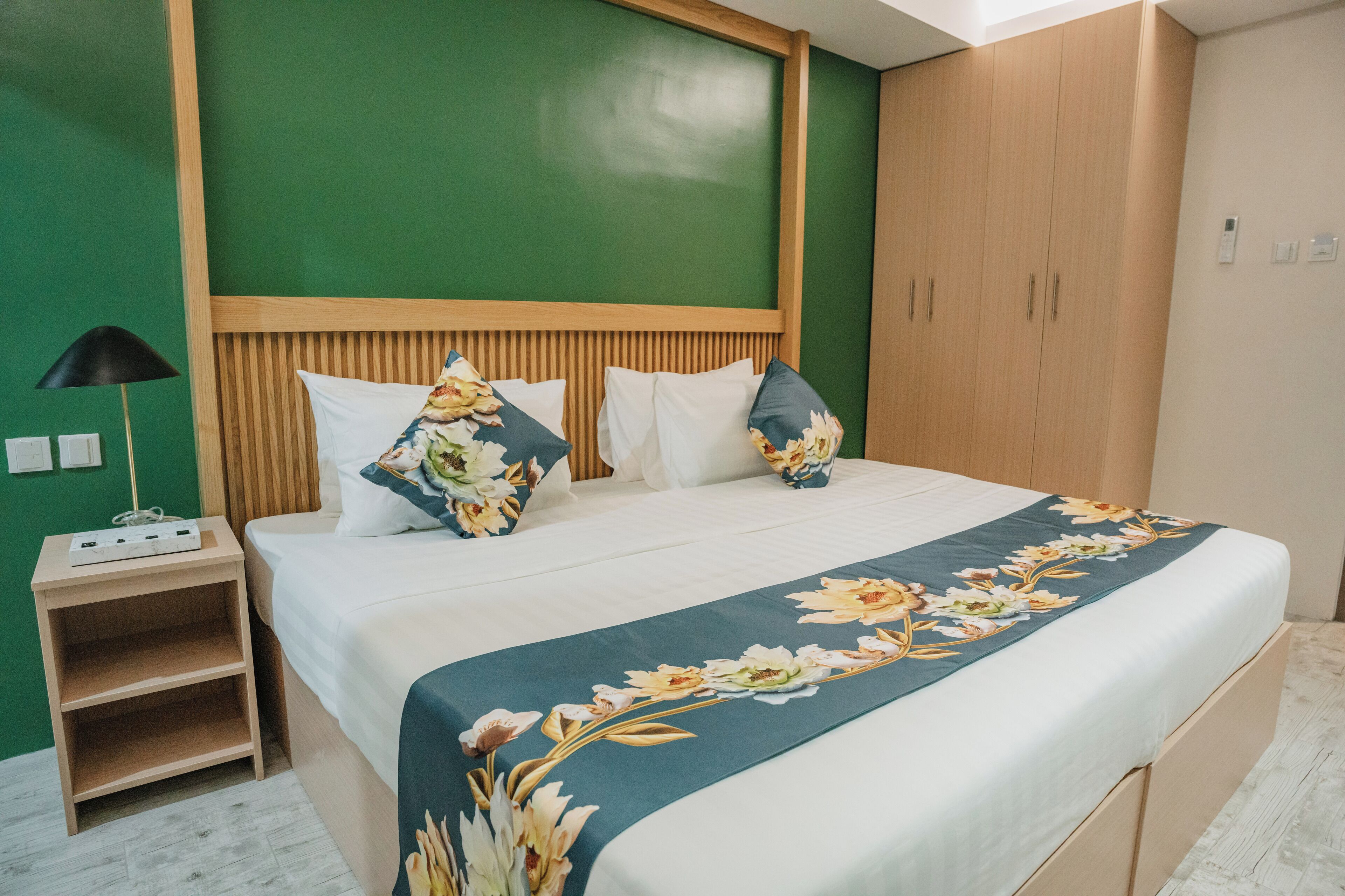 A neatly made bed with a floral bedspread, a bedside table with a lamp, and a wardrobe with a wooden finish.