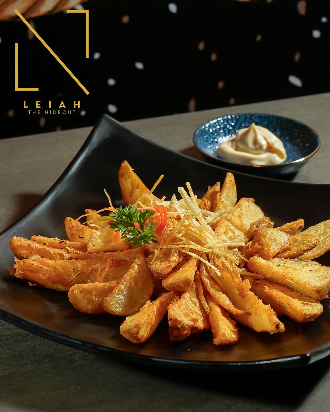 A plate of seasoned fries with a garnish, served on a dark plate with a small bowl of sauce in the background.