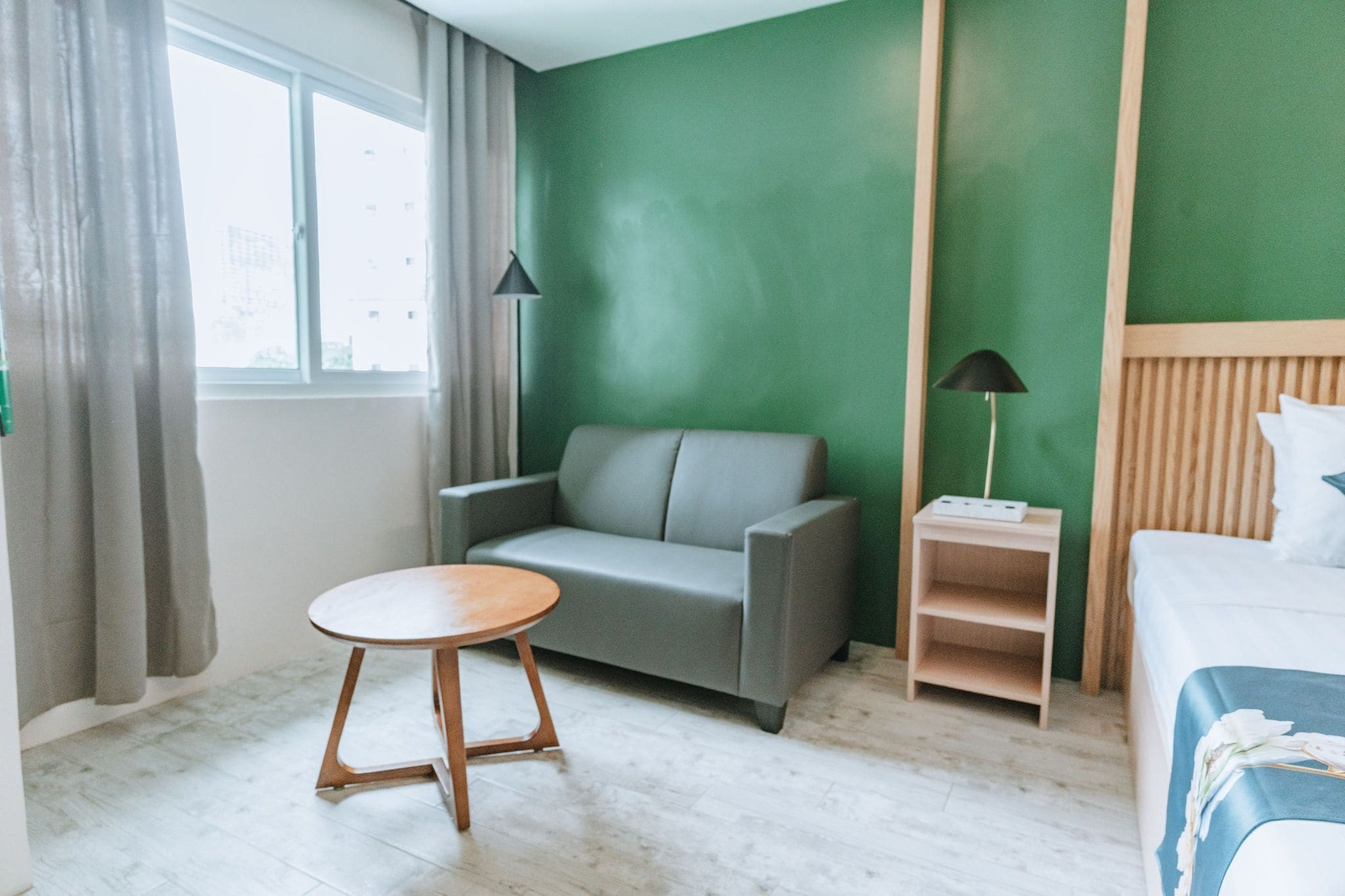 A modern hotel room with a grey sofa, a round wooden table, a bedside table with a lamp, and a bed with a blue cover.