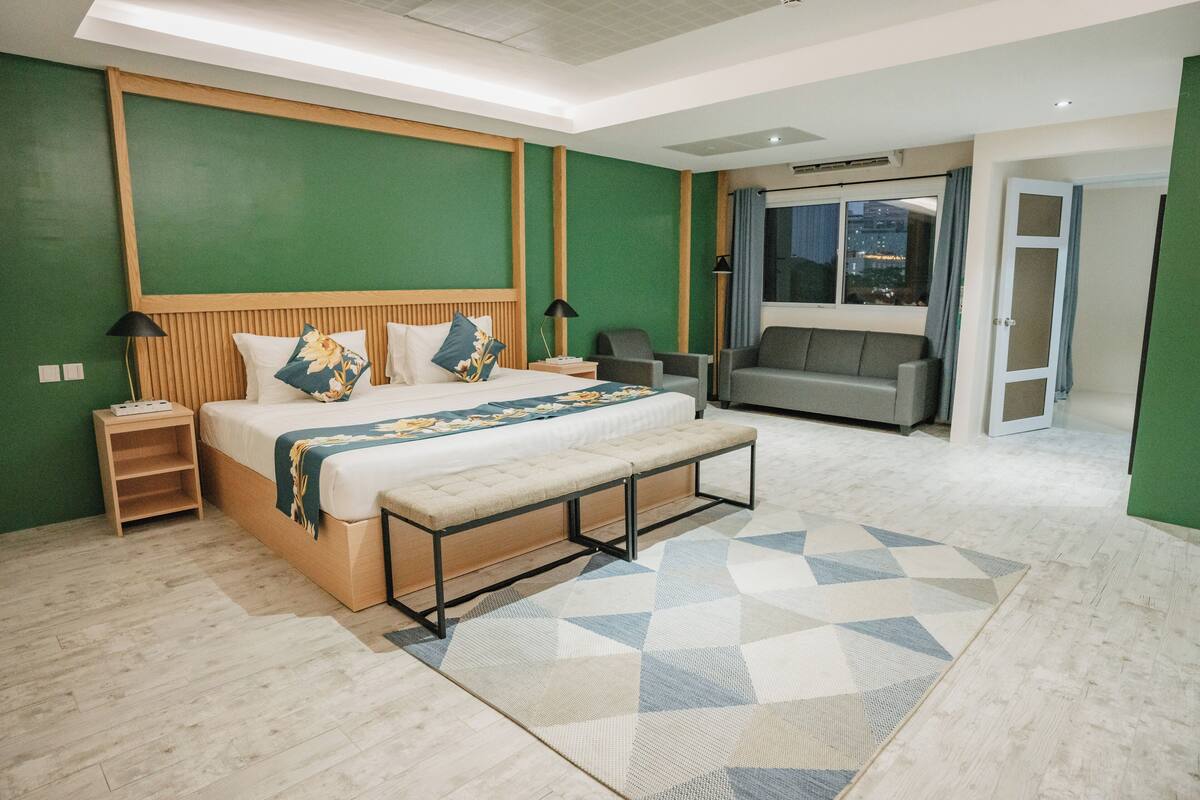 Una habitación de hotel moderna con una cama grande, una banca, un sofá y vistas a la ciudad a través del ventanal.