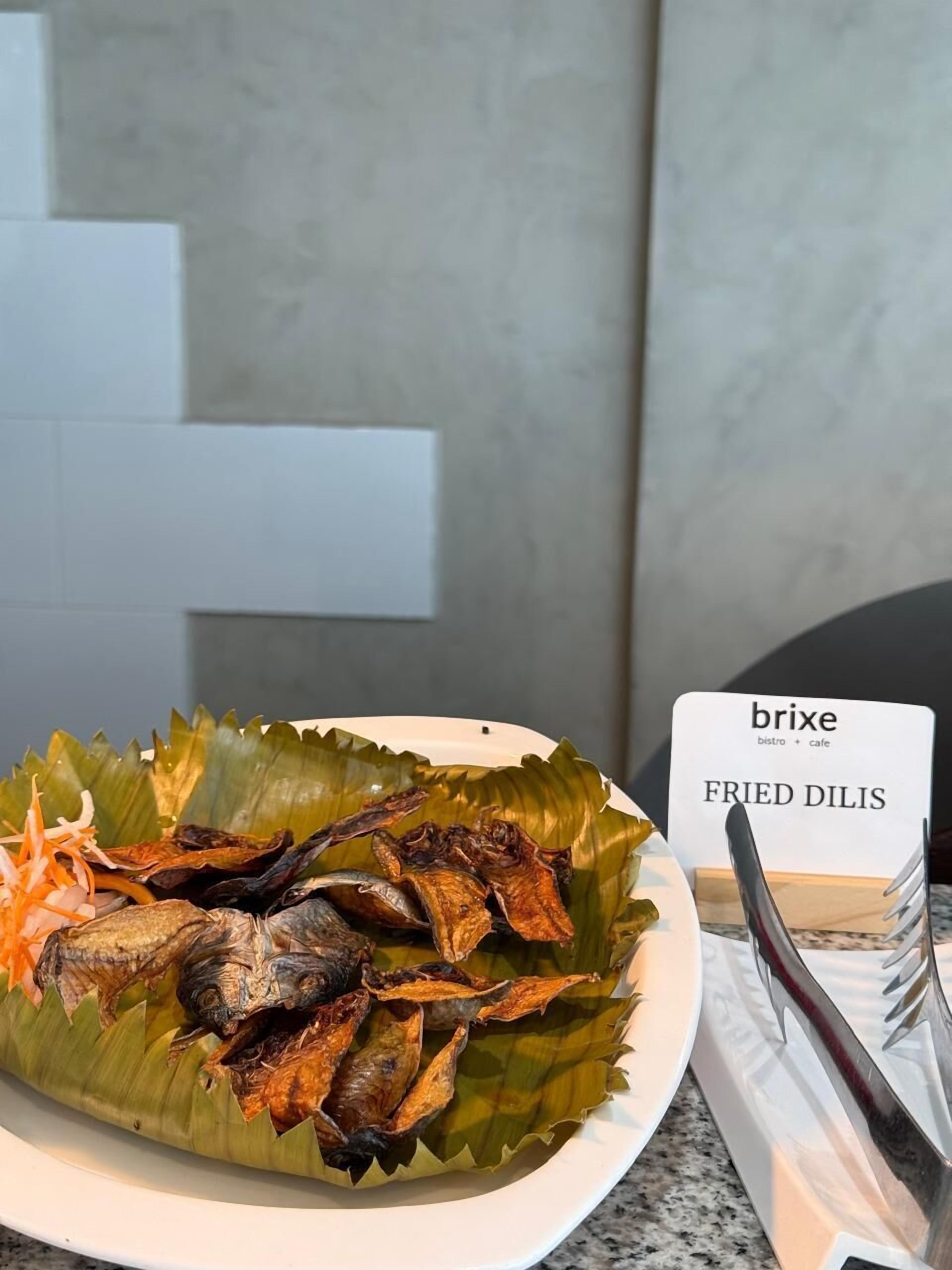 Un plato de pescado frito sobre una hoja de plátano con un letrero que dice 'FRIED DILIS' y una tarjeta de 'brixe bistro cafe'.