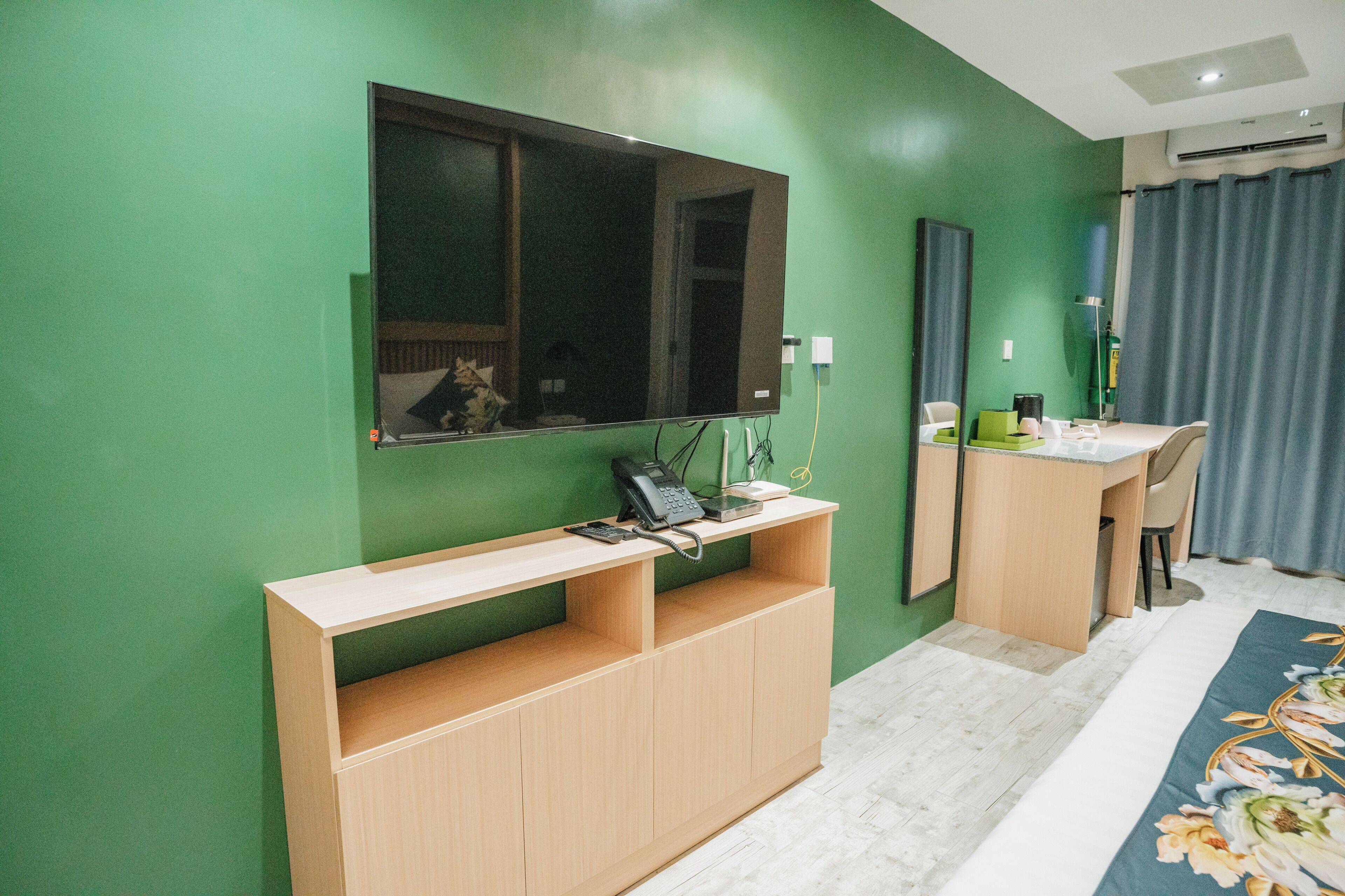 A room with a green wall, a wooden desk, a mirror, a bed with a floral bedspread, and a window with curtains.