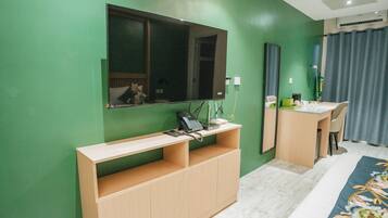 A room with a green wall, a wooden desk, a mirror, a bed with a floral bedspread, and a window with curtains.