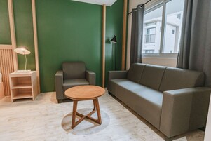 A modern living room with a green accent wall, a grey sofa, a wooden coffee table, and a small wooden side table with a lamp.