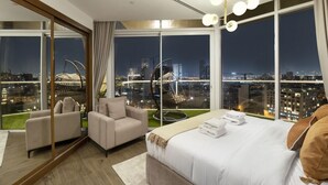 Une chambre d’hôtel moderne dotée d’un grand lit, d’un coin salon avec deux fauteuils et offrant une vue sur la ville grâce à de grandes fenêtres.