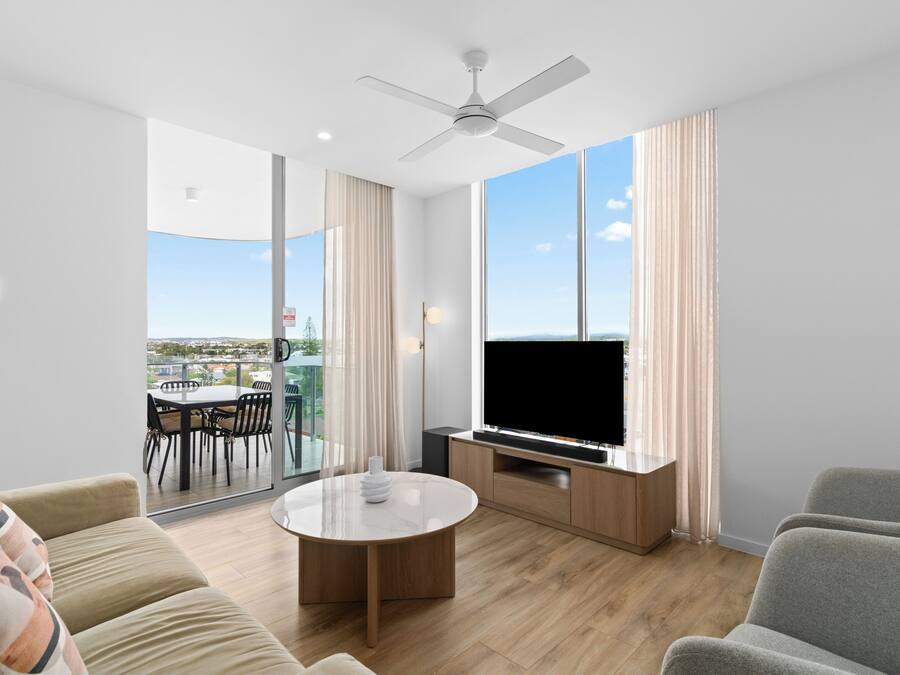 A modern living room with a flat-screen TV, a ceiling fan, a round coffee table, and a large window offering a view of the outdoors.