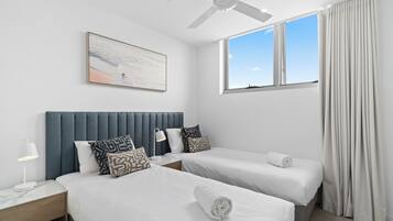A hotel room with two beds, a ceiling fan, a window with curtains, and a painting on the wall.