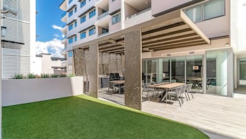 Un patio moderno al aire libre con zona de comedor, una pared blanca y un edificio con balcones al fondo.