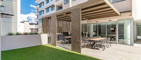 A modern outdoor patio with a dining area, a white wall, and a building with balconies in the background.