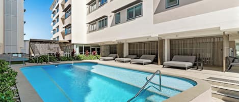 A swimming pool with lounge chairs, a covered seating area, and a building with multiple floors and balconies.