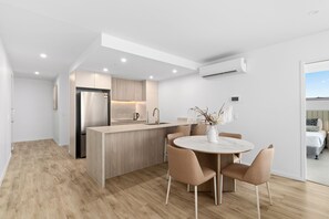 A modern kitchen with a central island, a dining area with a round table and chairs, and a bedroom visible through an open door.