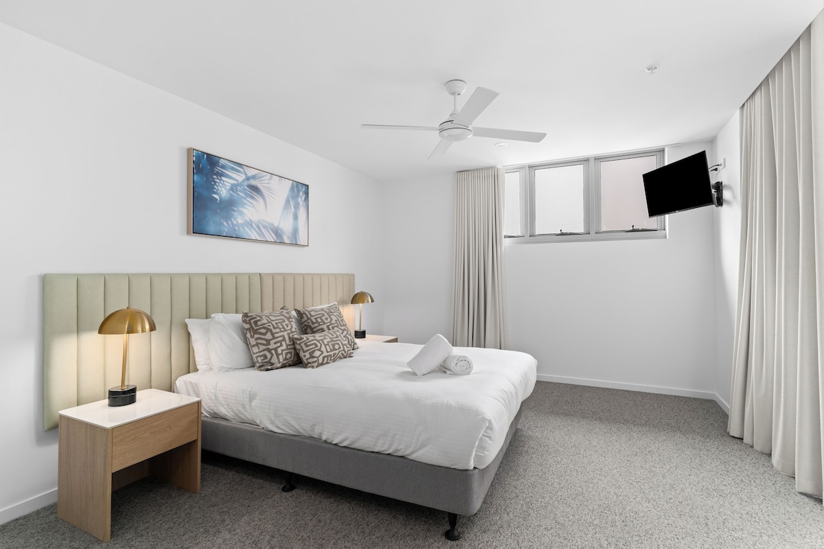 A modern hotel room with a bed, nightstands, a ceiling fan, a mounted TV, and a window with curtains.