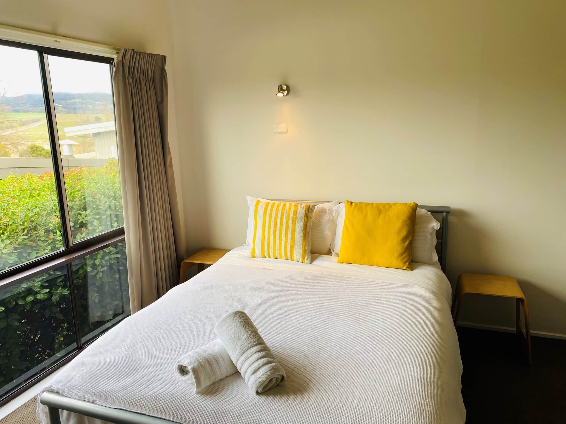A hotel room with a large window, a bed with white and yellow pillows, a rolled white towel, and two bedside tables.