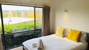 Habitación de hotel con un ventanal grande, cama con ropa blanca y almohadas amarillas, y vistas a zonas verdes y edificios.