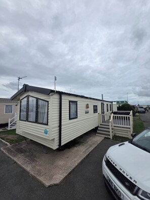 A mobile home with a ramp leading to the entrance, a vehicle parked nearby, and a cloudy sky.