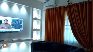 A living room with a dark blue sofa, a flat-screen TV mounted on the wall, and orange curtains.