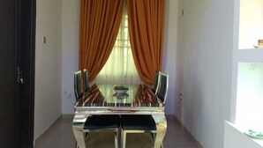 A dining area with a glass table and chairs, orange curtains, and a piano.