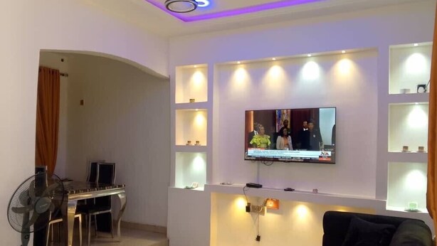 A modern living room with a flat-screen TV mounted on the wall, a black sofa, a coffee table, and a dining area with a table and chairs.