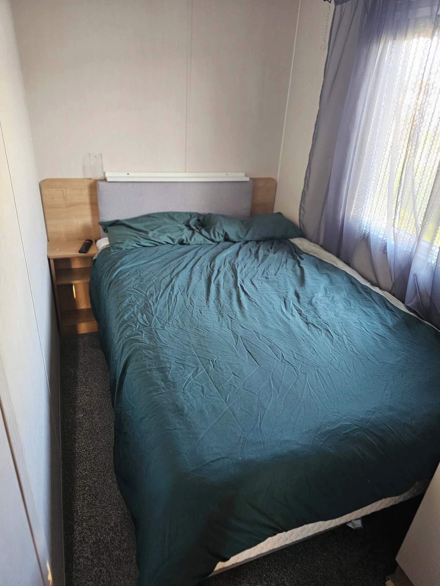 A small, compact room with a single bed, a wooden nightstand, and a window with sheer curtains.