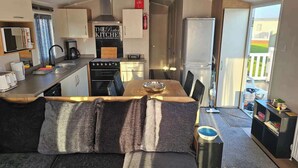 A compact kitchen with a stove, sink, and a refrigerator. There is a dining area with a table and chairs. The room has a sliding door leading to a balcony. - Luxury Chichester caravan  Seaside Retreat (Chichester)