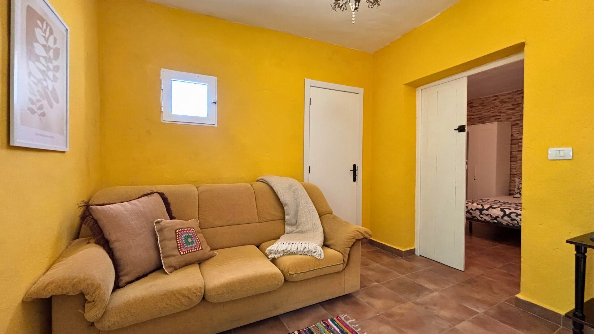 Una habitación con paredes amarillas, un sofá color beige, una puerta y una cama al fondo.