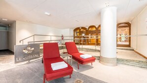 Una hall d'albergo con una reception, poltrone rosse, un grande pilastro e una scala che conduce a un piano superiore.