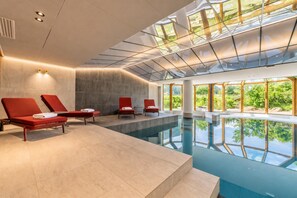 Zona de piscina interior moderna con sillones reclinables rojos, techo de cristal transparente y vistas al exterior con zonas verdes.