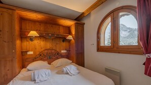 Una stanza con rivestimento in legno, un letto, due lampade a parete, una finestra con vista su colline innevate e un termosifone.