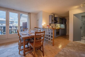Un comedor con mesa y sillas de madera, una cocina con armarios grises, y vistas a través de amplios ventanales.