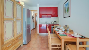 Een kleine keuken met rode kastjes, een houten eettafel met stoelen, en een koelkast.