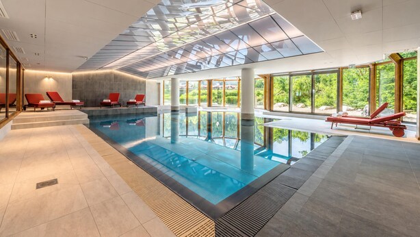 Piscina interior moderna con techo de vidrio transparente, sillas de descanso rojas y amplias ventanas que ofrecen vistas a zonas verdes.