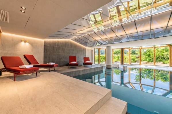 A modern indoor pool area with red lounge chairs, a clear glass ceiling, and a view of greenery outside.
